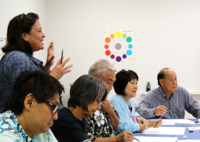 Photo of Meleanna Meyer standing up and gesturing using both hands while attendees sitting beside her are attentively listening