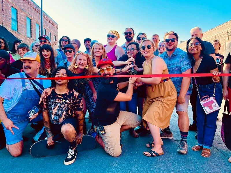 A smiling crowd of people outdoors, a few of them holding a large pair of scissors primed to cut a red ribbon.