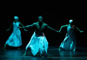 Three dancers in luminous white costumes gesture with open arms on a dimly lit stage.