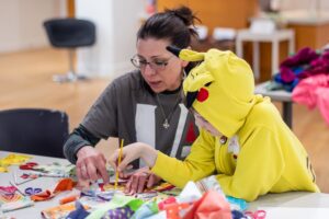 A teacher is helping a young student dressed in a yellow costume with painting in a classroom. On the table in front of them are colorful art supplies. 