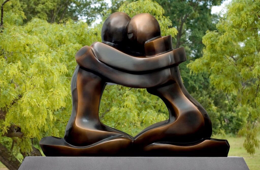 A outdoor sculpture in polished bronze portrays two stylized kneeling figures holding one another in an embrace.